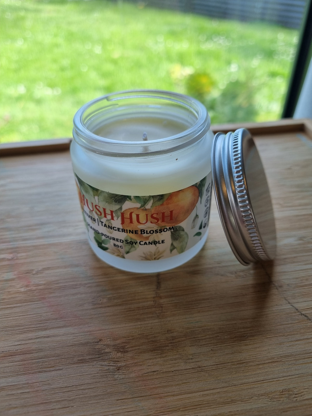 Opened jar soy candle in Hush Hush scent by Kirsche Candle Haus on a pine wooden tabletop, cream wax visible but candle is not lighted. Image background is green grass.