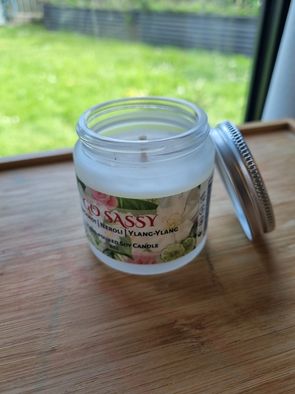 Kirsche Candle Haus hand-poured soy candle in Go Sassy scent sits on a cream tablecloth in a cozy living room setting. The silver lid is placed beside it on a small white dish, with pink rose petals and a book scattered nearby. Opened jar of Go Sassy artisan handpoured soy candle by Kirsche Candle Haus, made in Wellington NZ.