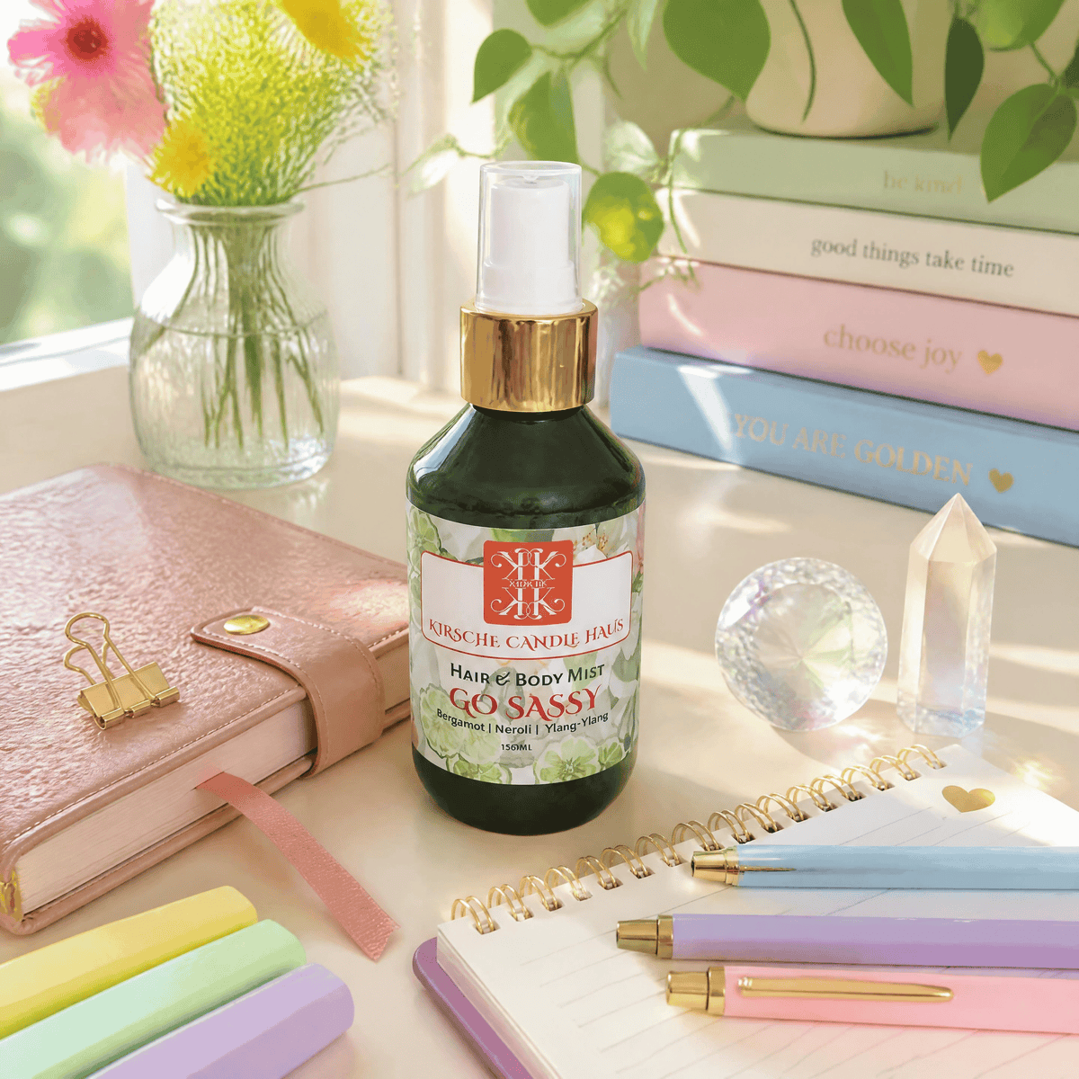 A bottle of Kirsche Candle Haus natural fragrance handmade in New Zealand Go Sassy Hair & Body Mist sits on a desk surrounded by stationery, books, and flowers in a bright, natural setting.