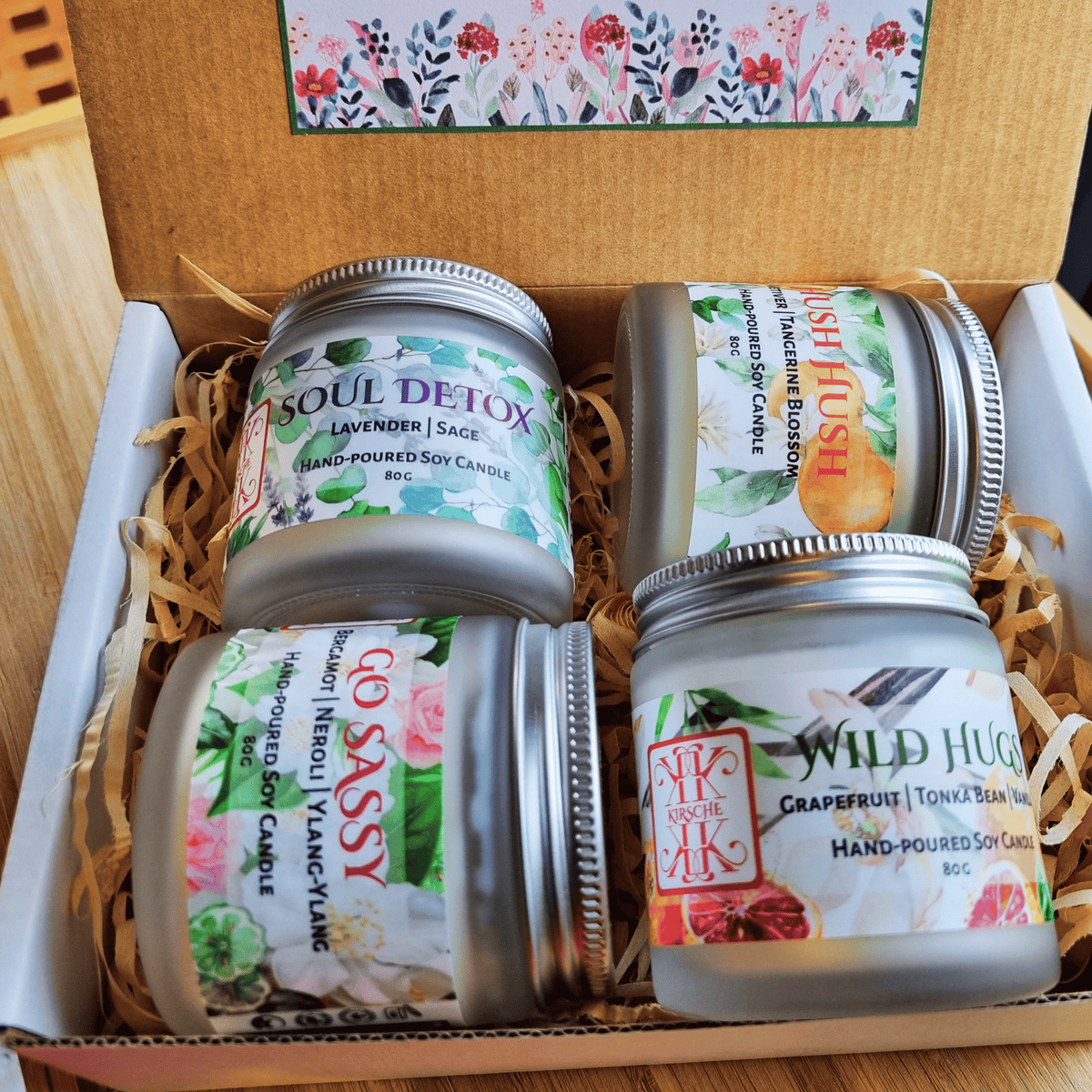Four Kirsche Candle Haus hand-poured soy candles in glass jars with silver lids, arranged in a cardboard gift box with paper shred. The labels feature floral designs and scents including Soul Detox, Hush Hush, Go Sassy, and Wild Hug. Small batch made in Wellington New Zealand.