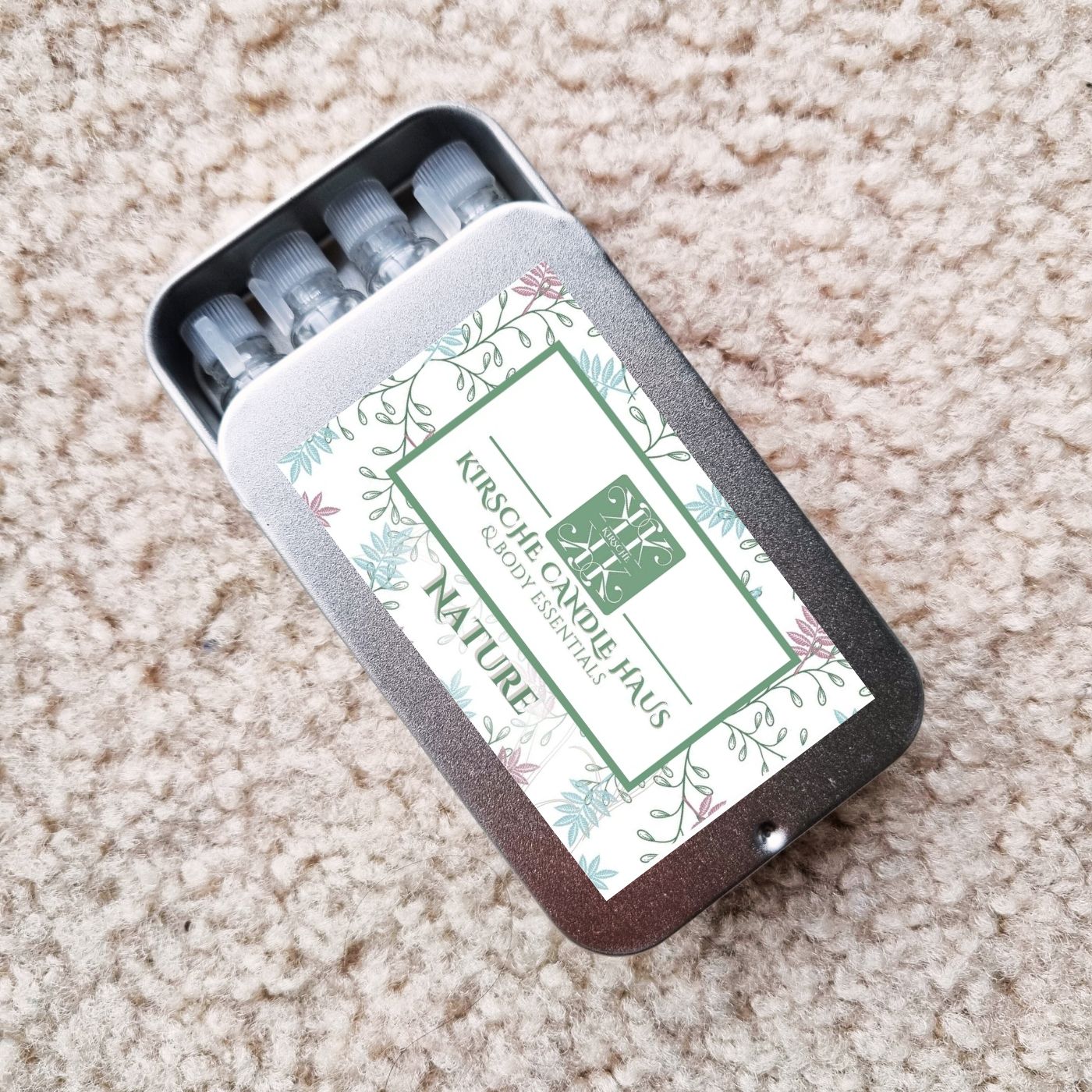 Set of small vials in a decorative tin labeled 'Kirsche Candle Haus' Nature; on a beige carpet.