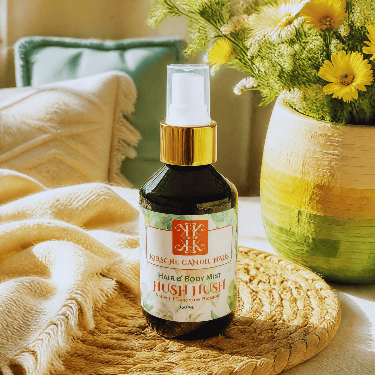 A bottle of handpoured NZ made  bodycare Kirsche Candle Haus Hair & Body Mist 'Hush Hush' fragrance, placed on a woven placemat next to a yellow and green striped vase of flowers.