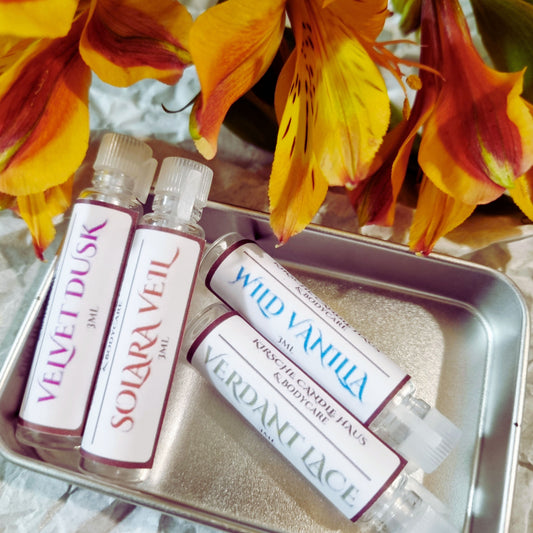 Four small vials of perfume labeled 'Velvet Dusk', 'Solara Veil', 'Wild Vanilla', and 'Verdant Lace' on a decorative tray with flowers in the background.