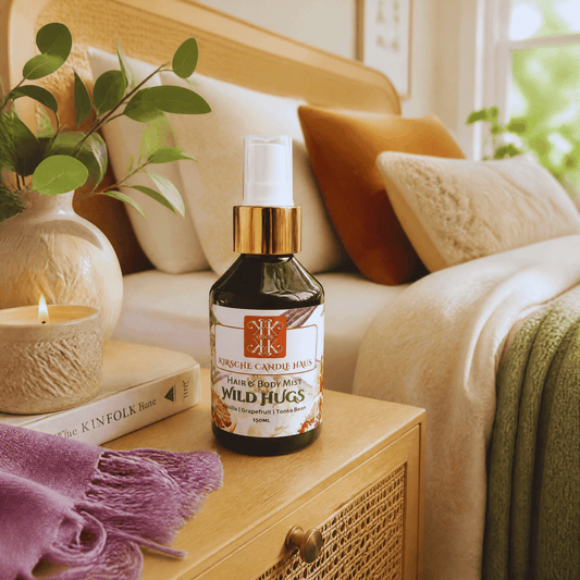 Kirsche Candle Haus 'Wild Hugs' hair & body mist spray bottle with golden cap sits on a wooden bedside table in a cozy bedroom setting, surrounded by a lit candle, a book, a purple throw blanket, and a bed with warm pillows.