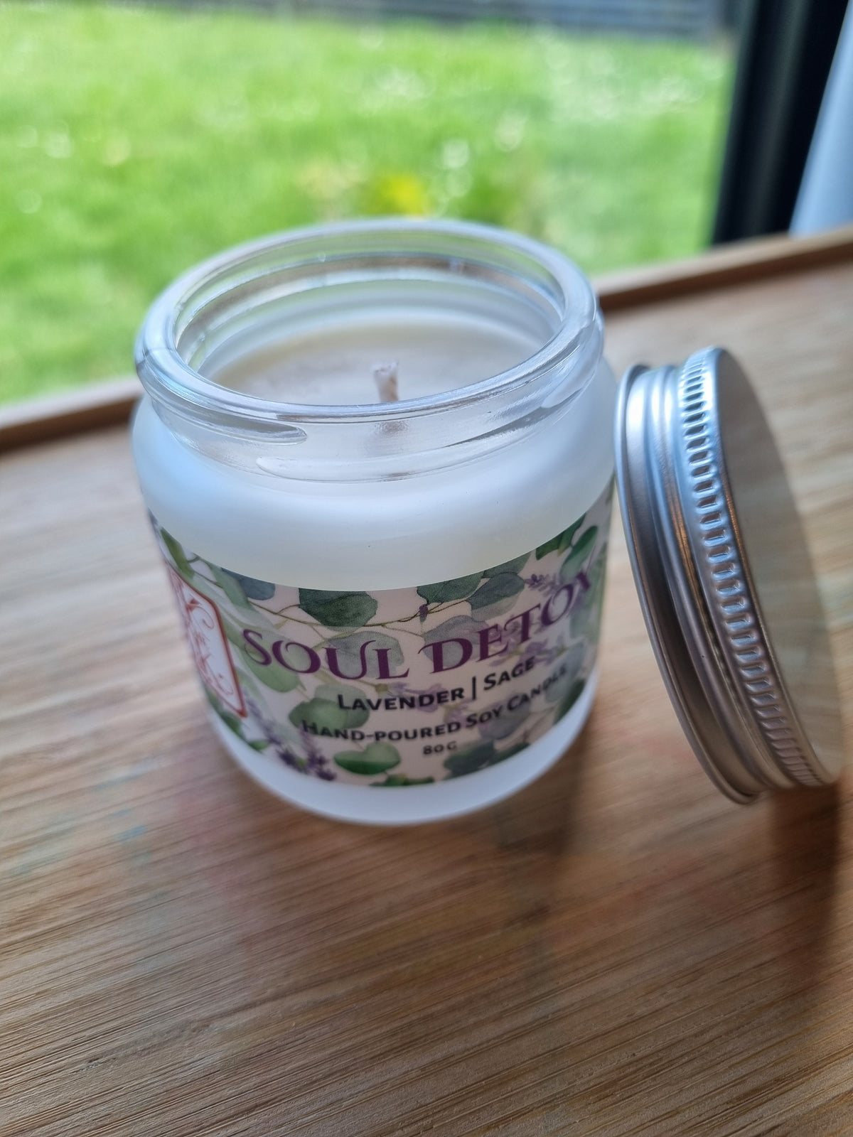 Kirsche Candle Haus artisan  soy candle labeled 'SOUL DETOX' sits on a wooden surface beside a window, lid opened and wax and wick visible but unlit.