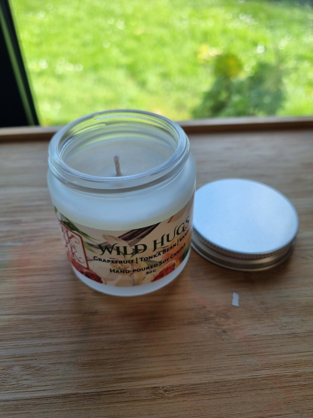 Kirsche Candle Hause Soy Candle jar of Wild Hugs Grapefruit, Tonka Bean, and Vanilla hand-poured soy candles sits on a wooden table in a cozy room setting near a window with lid open and candle wax and wick visible but unlighted. Handpoured in Wellington New Zealand.